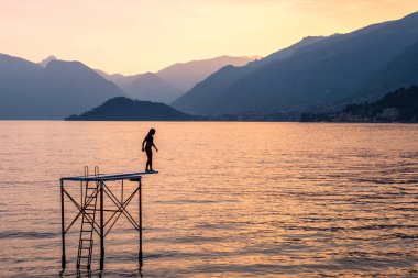 Suya platformdan atlamak çalışan bir kadın silueti. Gün batımında Dağları ile Como Gölü. Bulutlu gökyüzü. Bellagio, İtalya'dan görünüm