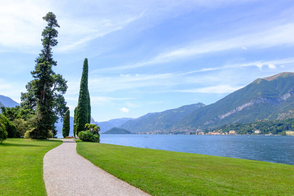 Road and grass field on shore of Lake Como. Mountains and cities on background. Bellagio, Italy