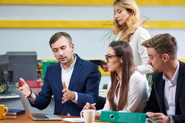 Team of mature women and men at meeting table discussing a business plan with laptop computer in office. Window on background