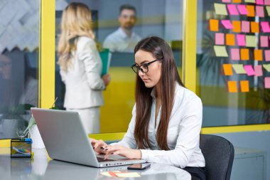 Genç iş kadını ofiste dizüstü bilgisayar üzerinde çalışıyor. İşçi ve arka plan üzerinde yapışkan notlar