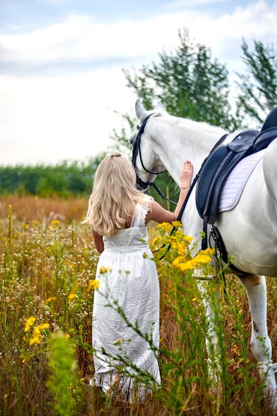 Chica en vestido blanco sosteniendo un caballo en el campo — Foto de Stock