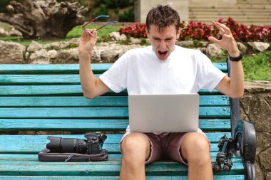 Kızgın genç fotoğrafçı parkta bir dizüstü bilgisayar üzerinde çalışıyor