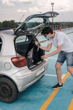 Sırt çantalı ve cerrahi maskeli genç bir adam arabadan havaalanına doğru yürüyor. Coronavirus pandemi salgını sırasında seyahat etmek