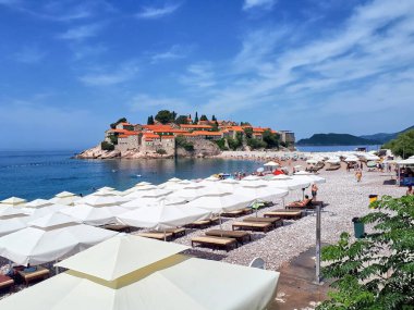 SVETİ STEFAN, MONTENEGRO - 9 Temmuz 2019: Yarımada, bir sürü şemsiyesi olan plaj ve ön planda dinlenen insanlar