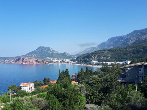 View of seafront of Sutomore with trees and buildings, beach with hills and mountains visible in the distance, Montenegro
