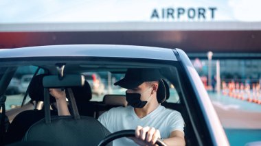 Tıbbi maskeli bir adam havaalanı yakınındaki bir arabada çantayla salgın sırasında güvenli bir yolculuğa çıkıyor.
