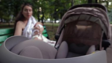 Arka planda bir bankta otururken bebeğini emziren bir anneyle bebek arabasının yakınlaşması.