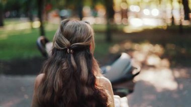 Uzun dalgalı saçlı bir anne bankta oturuyor, bebeğini huzurlu bir parkta emziriyor, modern bir bebek arabasının yanında.