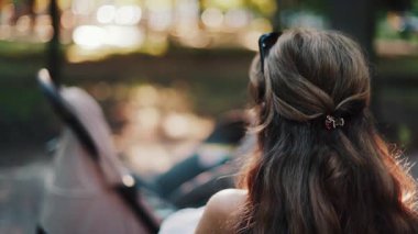 Uzun dalgalı saçlı bir anne bankta oturuyor, bebeğini huzurlu bir parkta emziriyor, modern bir bebek arabasının yanında.