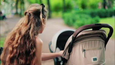 Yeşilliklerle çevrili bir bankta otururken bebek arabasını tutan kıvırcık saçlı bir kadın.