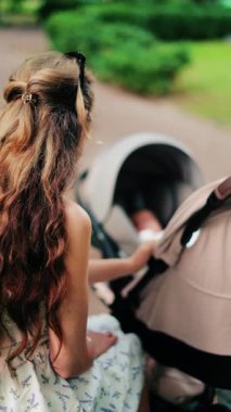 Yeşilliklerle çevrili bir bankta otururken bebek arabasını tutan uzun kıvırcık saçlı bir kadın. Dikey