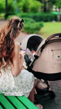 Yeşilliklerle çevrili bir bebek arabasının yanındaki bankta otururken telefonunda mesajlaşan uzun kıvırcık saçlı bir kadın. Dikey