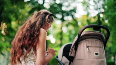 Yeşilliklerle çevrili bir bebek arabasının yanındaki bankta otururken telefonunda mesaj atan uzun kıvırcık saçlı bir kadın.