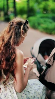 Yeşilliklerle çevrili bir bebek arabasının yanındaki bankta otururken telefonunda mesajlaşan uzun kıvırcık saçlı bir kadın. Dikey