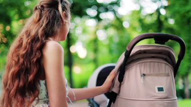 Yeşilliklerle çevrili bir bankta otururken bebek arabasını tutan kıvırcık saçlı bir kadın.