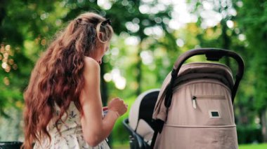 Yeşilliklerle çevrili bir bebek arabasının yanındaki bankta otururken telefonunda mesaj atan uzun kıvırcık saçlı bir kadın.