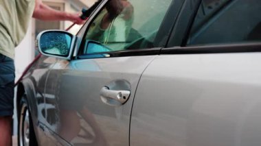 Chisinau, Moldova - 18 Ağustos 2025: Bir adam gümüş bir arabanın camını temizleme fırçasıyla yıkıyor, sabun ve kiri temizliyor