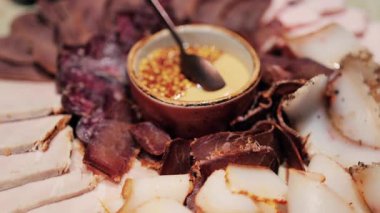 Şarküteri tahtasıyla çeşitli pişirilmiş etler, tütsülenmiş dilimler ve bir kâsede servis edilen hardal tohumu sosu.