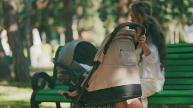 Genç anne parkta bir bebek arabasının yanında oturuyor, bebeğiyle dışarıda huzurlu bir anın tadını çıkarıyor.