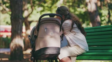 Genç anne parkta bir bebek arabasının yanında oturuyor, bebeğiyle dışarıda huzurlu bir anın tadını çıkarıyor.