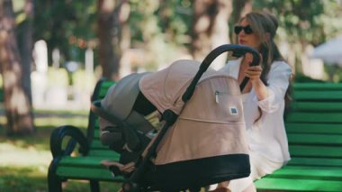 Genç anne parkta bir bebek arabasının yanında oturuyor, bebeğiyle dışarıda huzurlu bir anın tadını çıkarıyor.
