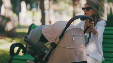 Anne parkta yeşil bir bankta oturuyor, bebeği içindeyken bebek arabasını nazikçe sallıyor.