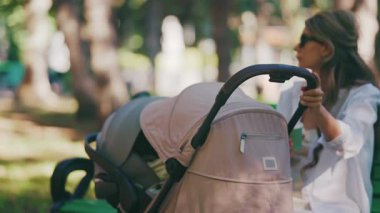 Genç anne parkta bir bebek arabasının yanında oturuyor, bebeğiyle dışarıda huzurlu bir anın tadını çıkarıyor.