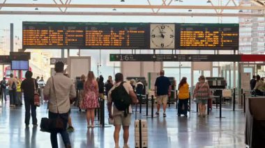 Alicante, İspanya - 19 Mayıs 2025: Valizli yolcular, büyük kalkış tahtaları ve analog bir saatin altındaki parlak bir tren istasyonunda hareket ediyorlar