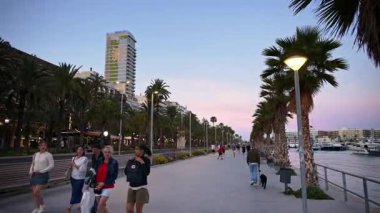 Alicante, İspanya - 26 Mayıs 2025: Gezginler gün batımında sallanan yatlarla palmiye püsküllü iskeleyi paylaşıyorlar