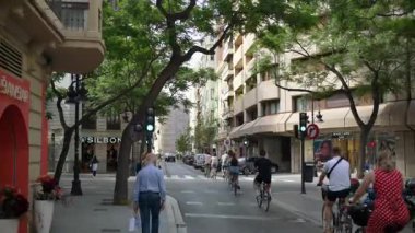 Valencia, Spain - May 28, 2025: Commuters and shoppers ride bikes through a green light on a tree lined avenue flanked by boutiques and mid rise residences