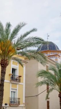 The iconic blue-tiled dome of Iglesia de San Jaime y Santa Ana framed by palm fronds and pastel facades in the Old Town of Benidorm, Costa Blanca, Spain. Vertical