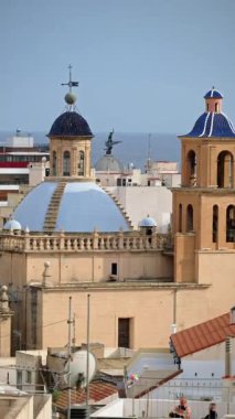 Narrow old-town street frames a blue-tiled church dome in Alicante, Spain. Vertical