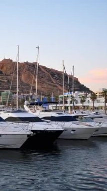 Alicante, Spain - May 26, 2025: A line of luxury boats rests with the Melia Alicante as a backdrop in soft pink skies. Vertical