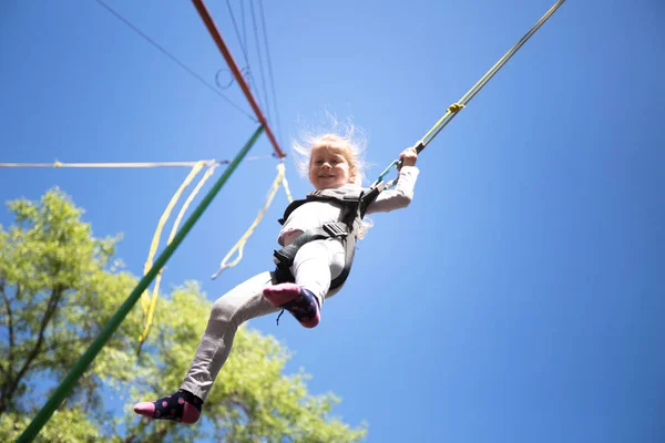 Child get fun on children trampoline with safety ropes. Little girl jumping high on blue sky background. Children summer entertainment concept.