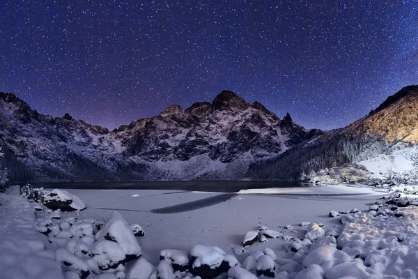 Winter night. Starry sky above mountain peak. Christmas night background. Winter wonderland. Winter landscape with ice covered lake at mountains and clear deep blue night sky full of stars.
