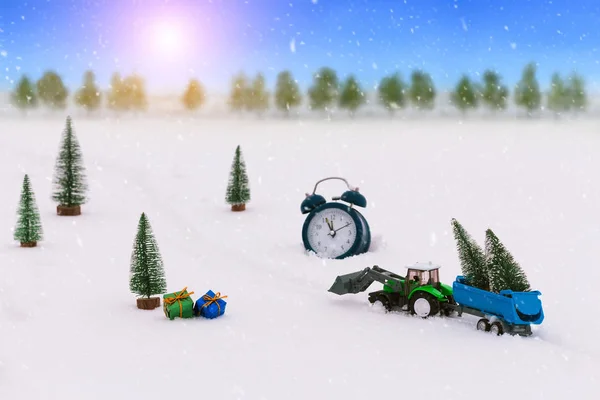 Toy tractor with a trailer carries Christmas trees during snowfall, rides through the snow in the middle of the forest. Beautiful background for greeting card. Winter composition. Happy holiday mood. Wake up, Xmas is coming.