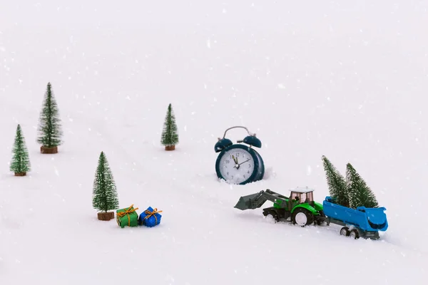 Toy tractor with a trailer carries Christmas trees during snowfall, rides through the snow in the middle of the forest. Beautiful background for greeting card. Winter composition. Happy holiday mood. Wake up, Xmas is coming.