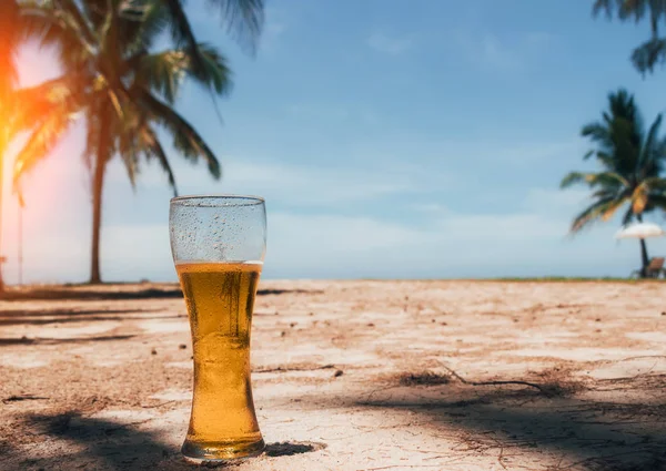 Misted glass of cold beer on the sand at the background of green palm trees, blue sky and sea coast. Tropical island. Heavenly delight.