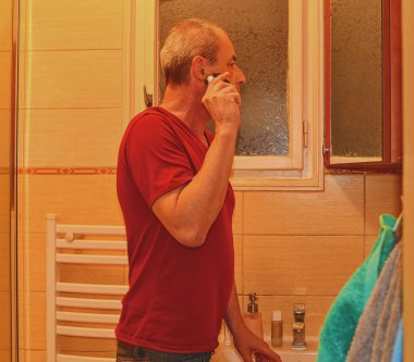 Orta yaşlı adam bir banyoda tıraş, elektrikli jilet kullanarak. Kıdemli konsept. Evde olgun adam.