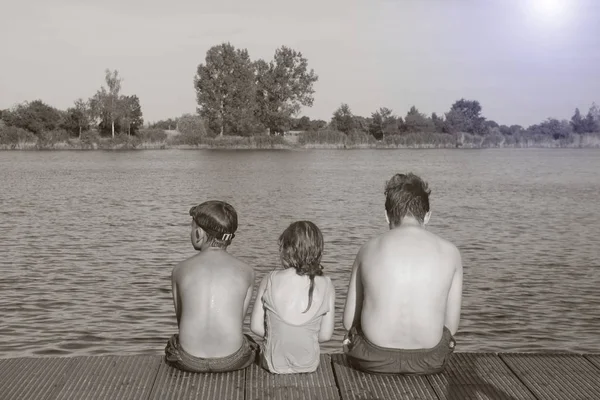 Gyermekek, ül a mólón. Három gyermek különböző kor - tinédzser fiú, fiú elemi kor és óvodai lány ül egy fa mólón. Nyári és a gyermekkor fogalma. Gyermekek a pad a tó — Stock Fotó