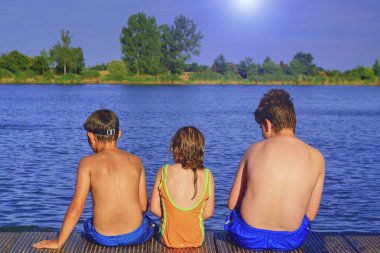 İskelede oturan çocuk. Üç farklı yaş çocuklar - genç çocuk, İlköğretim yaş çocuk ve ahşap bir iskele üzerinde oturan okul öncesi kız. Yaz ve çocukluk kavramı. Çocuk bankta göle