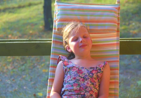 La bambina è seduta su una sdraio da giardino su una veranda. La bambina si sta prendendo un bagno di sole in giardino. Immagine sognante e romantica. Estate e felice concetto di infanzia — Foto Stock