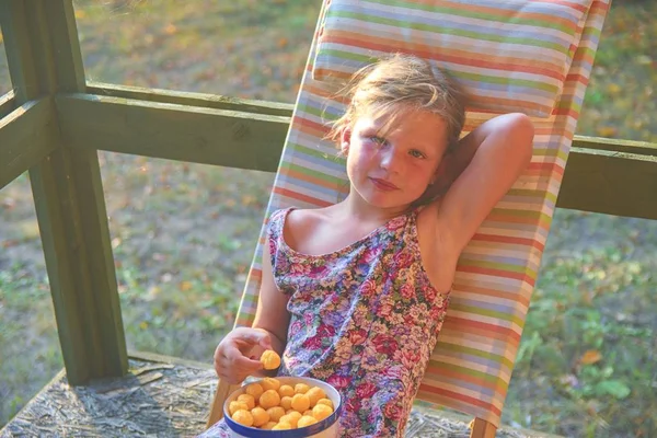 La bambina è seduta su una sdraio da giardino su una veranda. La bambina sta mangiando snack aromatizzati al formaggio in giardino. Immagine sognante e romantica. Estate e felice concetto di infanzia — Foto Stock