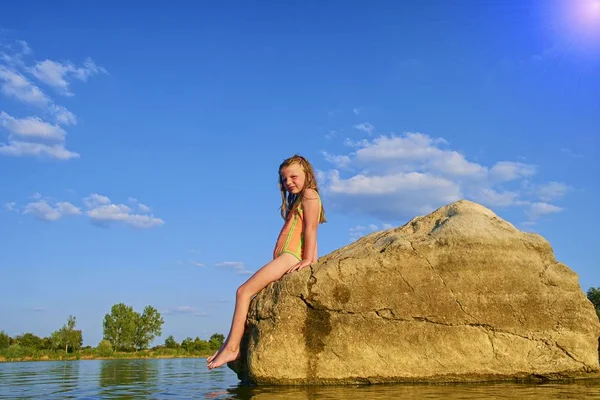 Portrait of child blond girl posing in swimsuit on rocks inside the lake at sunset. Summer and happy childhood concept. Copy space in bright blue sky — Stock Photo, Image