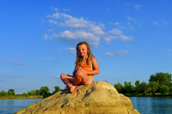 La bambina in costume da bagno si siede sulla roccia e guarda in lontananza. Estate e felice concetto di infanzia. Copia lo spazio nel cielo blu brillante — Foto Stock