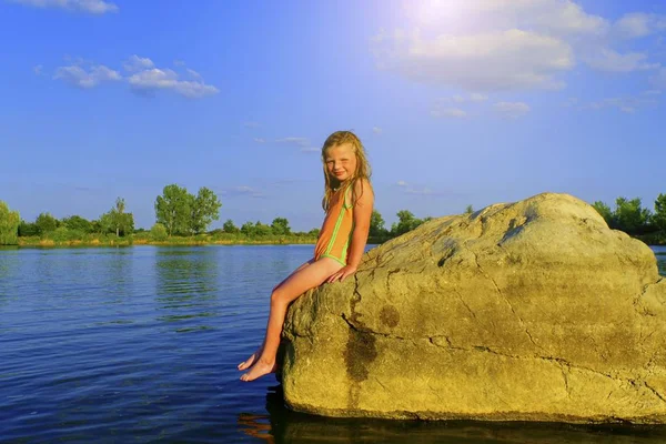 Cute little girl in a bathing suit sitting on a large rock by the lake at sunset. Summer and happy childhood concept. Copy space in bright blue sky — Stock Photo, Image