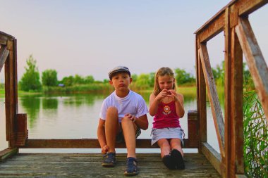 İskelede oturan çocuk. İki farklı yaş - İlköğretim çocuklarının ahşap bir iskele üzerinde oturan kız çocuk ve okul öncesi yaş. Kız kalp şekil verme. Yaz ve çocukluk kavramı. Çocuk bankta göle.