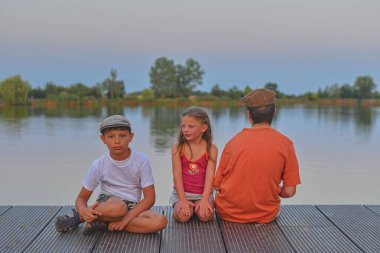 İskelede oturan çocuk. Kardeşler. Üç farklı yaş çocuklar - genç çocuk, İlköğretim yaş çocuk ve ahşap bir iskele üzerinde oturan okul öncesi kız. Yaz ve çocukluk kavramı. Çocuklar göle bankta. Tatil akşam sırasında altın saat. 