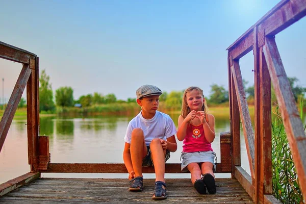 İskelede oturan çocuk. İki farklı yaş - İlköğretim çocuklarının ahşap bir iskele üzerinde oturan kız çocuk ve okul öncesi yaş. Kız kalp şekil verme. Yaz ve çocukluk kavramı. Çocuk bankta göle.