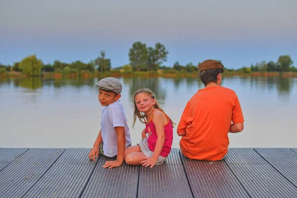 Children sitting on pier. Siblings. Three children of different age - teenager boy, elementary age boy and preschool girl sitting on a wooden pier. Summer and childhood concept. Children on bench at the lake. Golden hour during vacation evening.